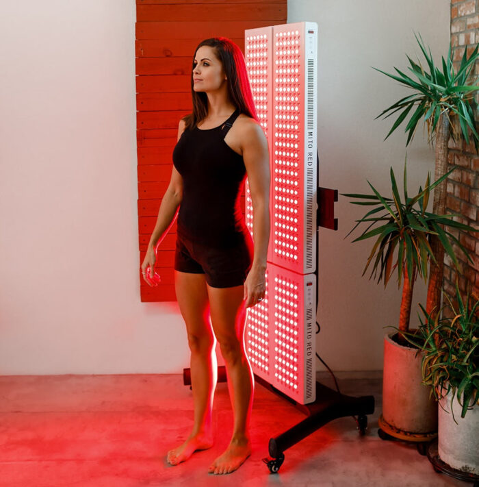 Young woman in black bathing suit standing in front of red light therapy panel hung on wall with plant behind her