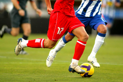 Soccer player in red running into a yellow soccer ball with referee behind him